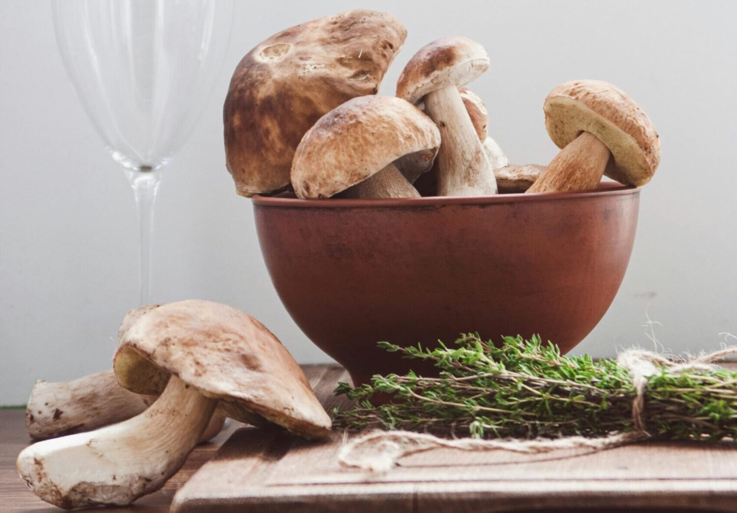 Fresh porcini mushrooms in a bowl beside herbs and a wine glass, ready for a pasta recipe.