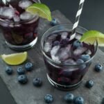 Two glasses of blackberry sangria garnished with fresh blueberries, lime wedges, and striped straws—served on a slate board for a vibrant and inviting drink presentation.