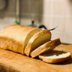 Freshly baked Ezekiel bread loaf partially sliced on a wooden cutting board, showcasing its golden crust and airy texture.