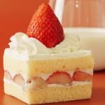 Slice of strawberry shortcake with whipped cream and fresh strawberries, served on a warm-toned surface beside a glass of milk.