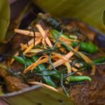 Close-up of beef giniling randang served in a clay pot lined with banana leaves, topped with fried vegetables and green chili peppers.
