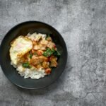 A bowl of steamed rice topped with crispy fried egg, stir-fried pork, chopped red chilies, and fresh greens, capturing a homemade take on Pepper Lunch.