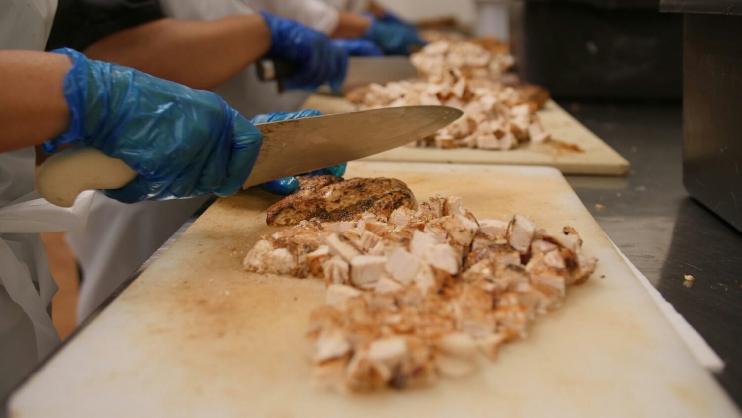 Close-up of gloved hands chopping grilled chicken breast into cubes on a white cutting board in a commercial kitchen, preparing ingredients for Chicken Marsala.