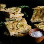 Slices of golden-baked Italian scarpaccia topped with lemon slices and rosemary, arranged with a small wooden bowl of flaky sea salt.