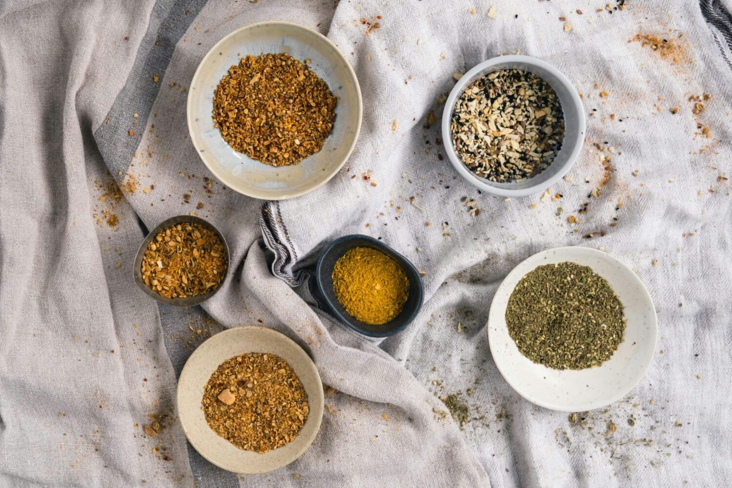 Assorted dried herbs and spices in small bowls on a cloth, ready to mix into a homemade remedy.