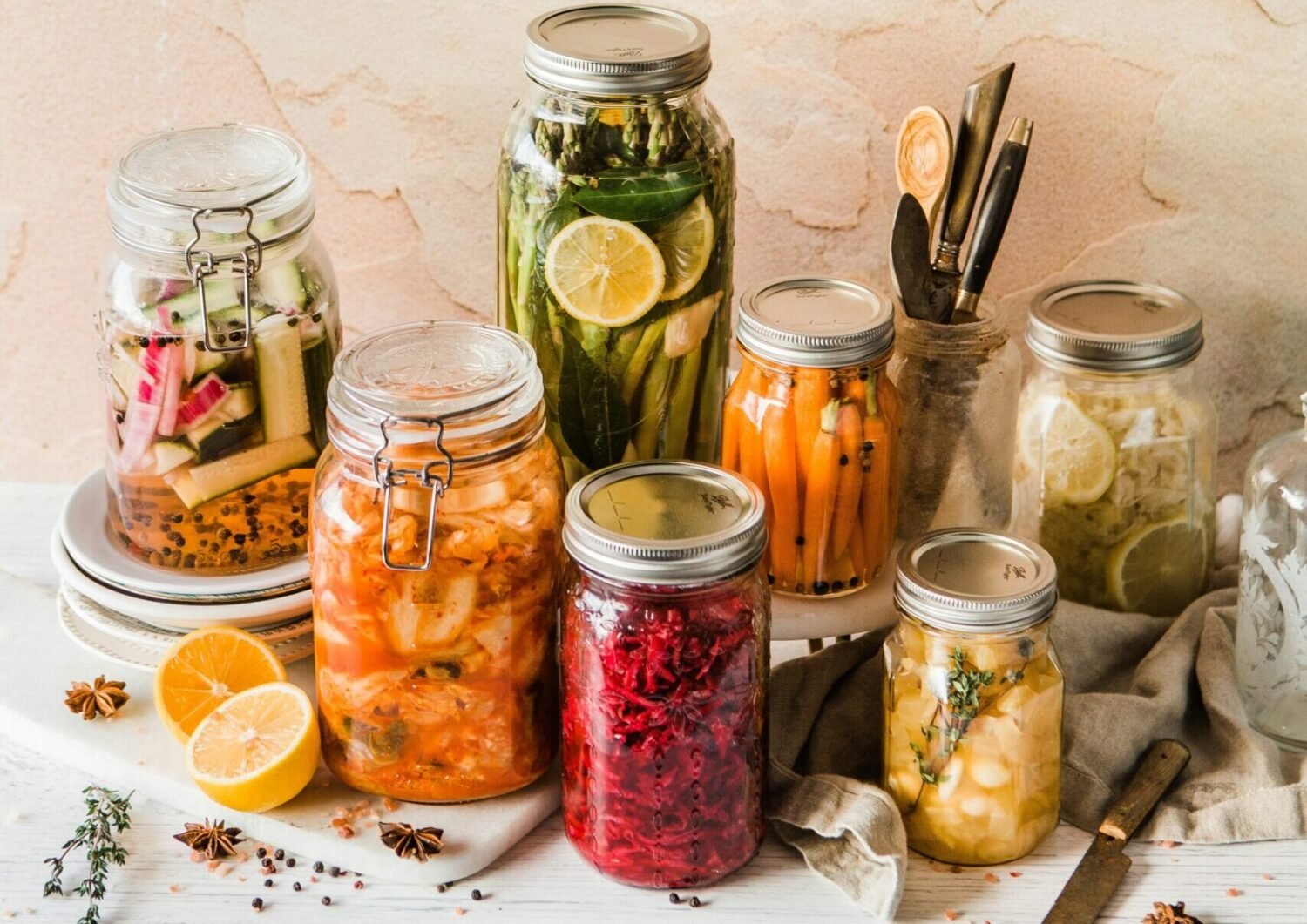 Assorted jars of homemade pickled and preserved vegetables and fruits arranged on a white table with rustic decor.